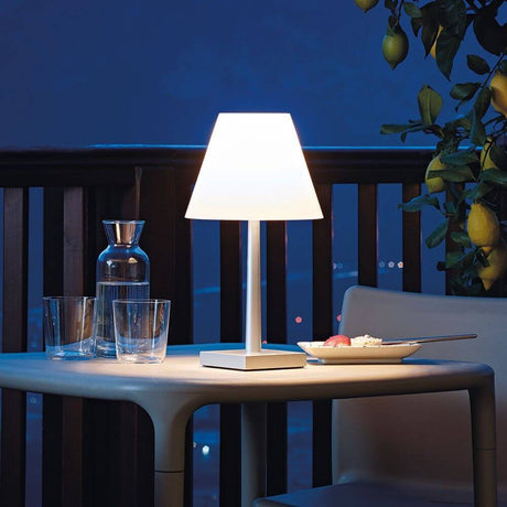 Rotaliana Dina+ battery-operated LED table lamp with charging station, white, on an outdoor table at night.