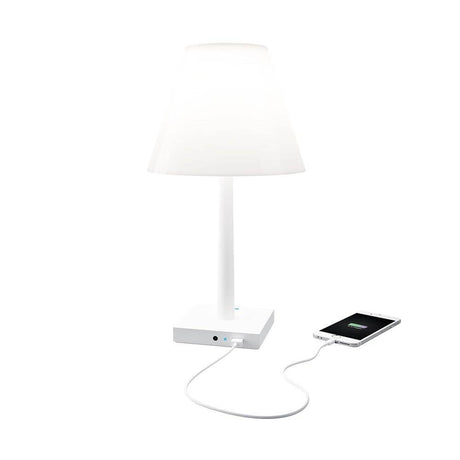 Rotaliana Dina+ battery-operated LED table lamp with charging station, white, on a table with a smartphone charging cable.