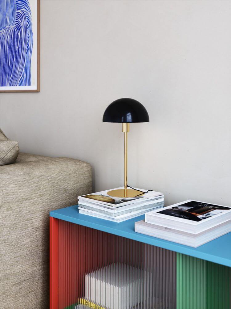 Ellen 20 Tischlampe auf einem Tisch mit buntem Regal und Literatur im Hintergrund.