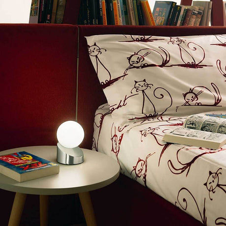 Gravity LED Tischleuchte aus Metall & Glas auf einem Nachttisch neben einem Bett mit Katzenbettwäsche.