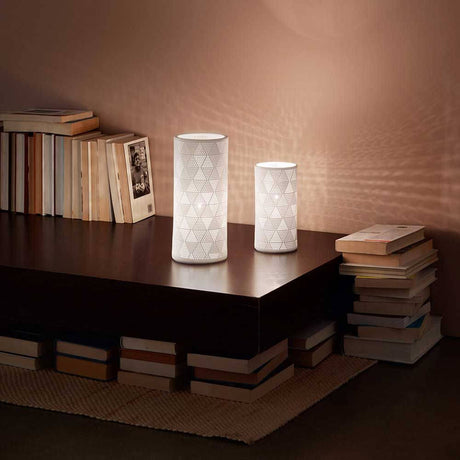 Tischleuchte Micol in Weiß auf einem Tisch mit Bücherstapeln und sanftem Licht im Hintergrund.