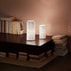 Tischleuchte Micol in Weiß auf einem Tisch mit Bücherstapeln und sanftem Licht im Hintergrund.