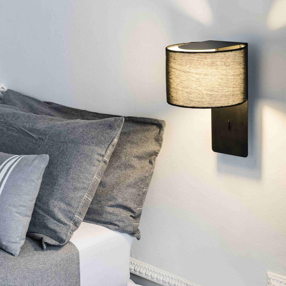 Fold Wandleuchte neben einem Bett mit grauen Kissen, modernes Design mit Stoffschirm und integriertem Schalter.