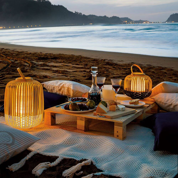 Lampe de sol à batterie Forestier LED Take A Way IP66 sur une table de plage avec le crépuscule et des boissons.