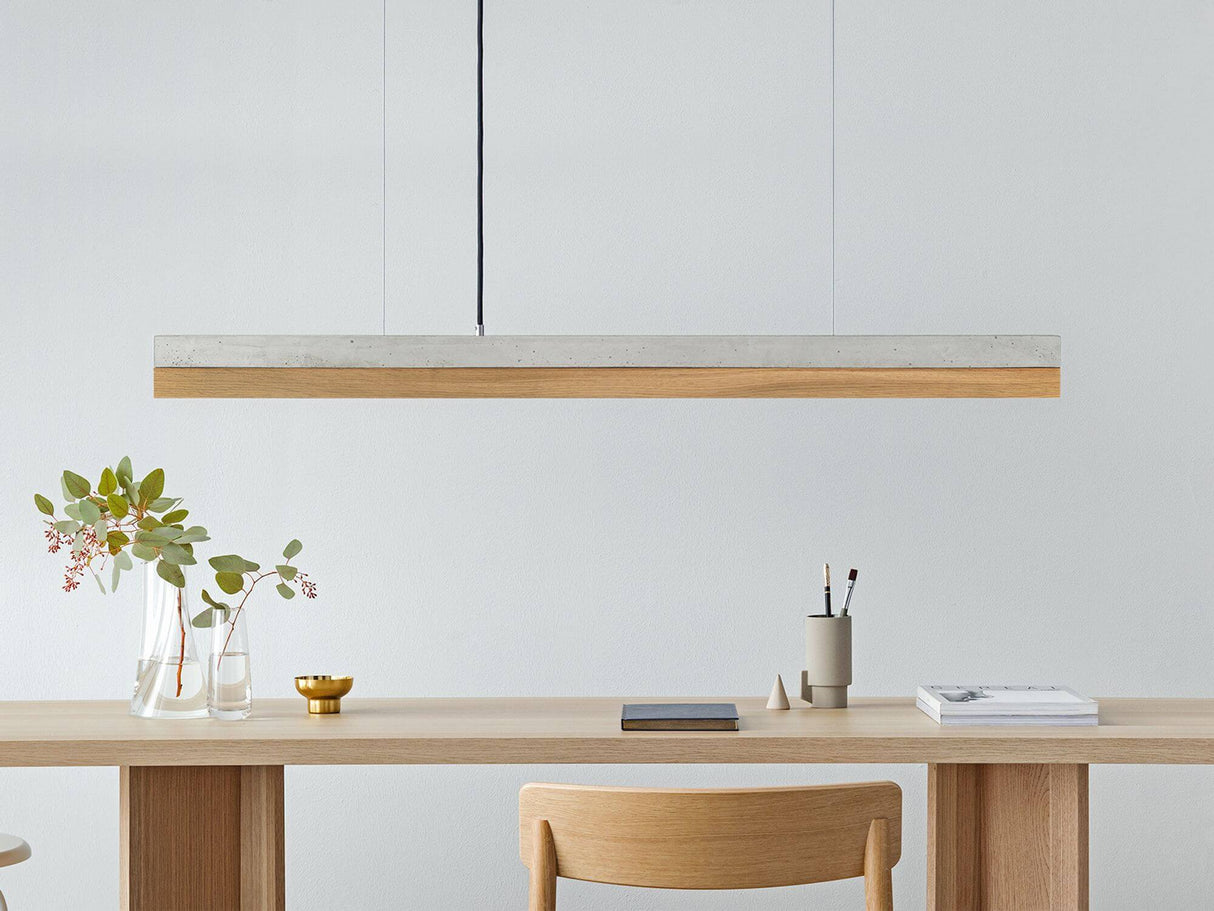 Gantlights C1 LED-Pendelleuchte 122cm Beton Holz über einem modernen Tisch mit Holzstuhl und Pflanzen.