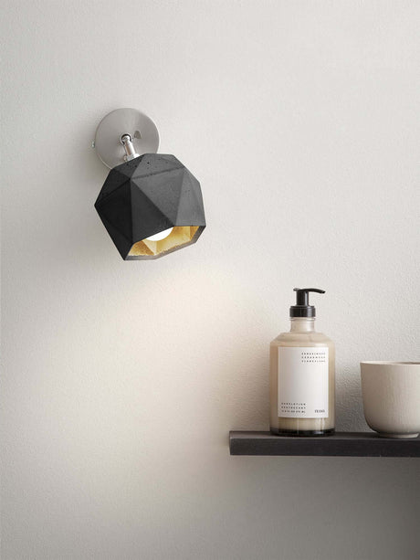 Gantlights T2 Wandspot Beton trianguliert an grauer Wand mit warmem Licht und Pflegeprodukt auf Regal