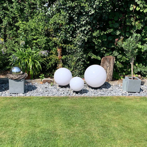Dekorative weiße Kugeln und Pflanzen im Garten, umgeben von Steinen und modernen Pflanzkästen.
