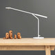 Grazile LED Tischleuchte Flow 5W in Weiß auf einem Holztisch mit dekorativen Holzfiguren im Hintergrund.