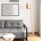 Holz Stehlampe mit goldenem zylindrischem Schirm neben grauem Sofa in modernem Wohnambiente.