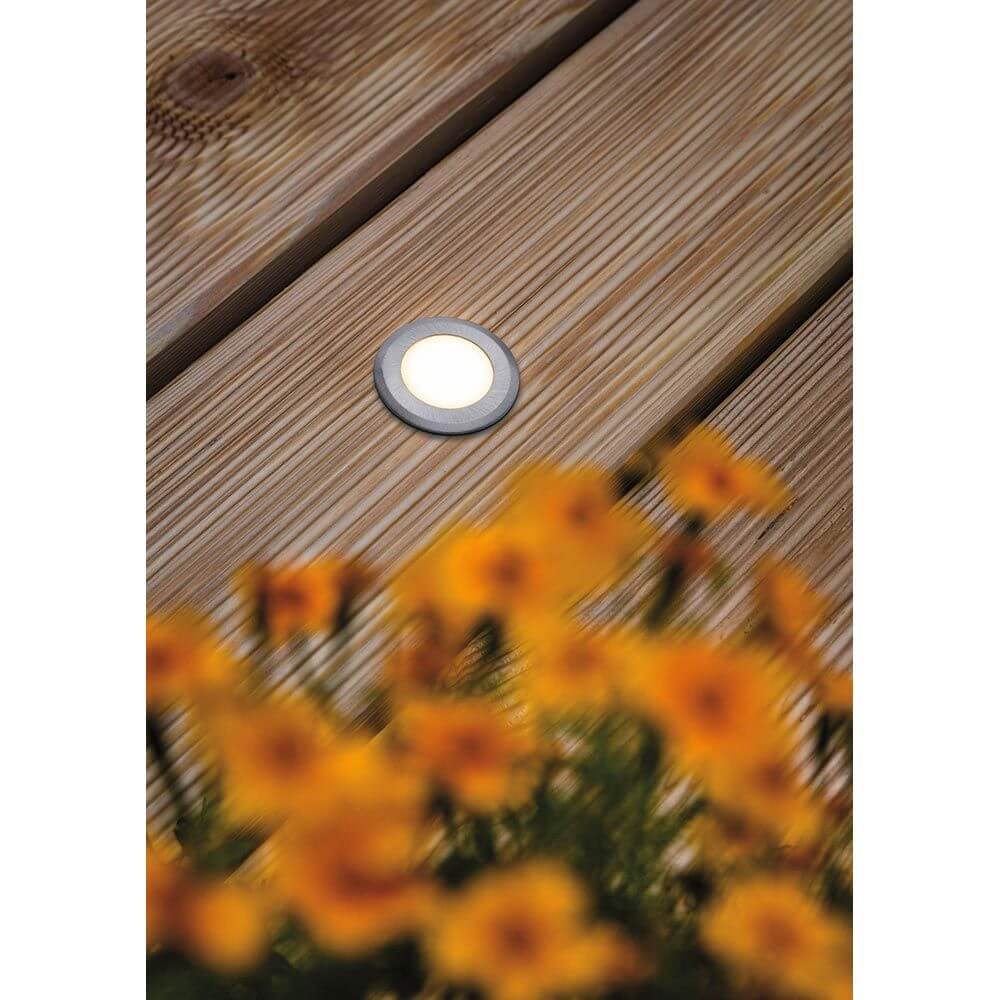 House LED Außen Bodeneinbauleuchte Rund Edelstahl IP67 auf Holzdeck mit orangefarbenen Blumen im Vordergrund.