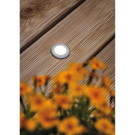 House LED Außen Bodeneinbauleuchte Rund Edelstahl IP67 auf Holzdeck mit orangefarbenen Blumen im Vordergrund.