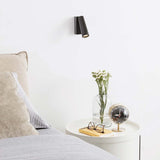 Pipe LED Wandleuchte Strahler in modernem Schlafzimmer neben Tisch mit Blume und Uhr.