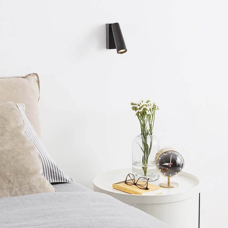 Pipe LED Wandleuchte Strahler über einem Nachttisch mit Blumen und einer Tischuhr.