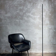 Yoko LED lampadaire en métal à côté d’une chaise noire moderne devant un mur gris.