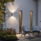 Imola LED Außen-Wandleuchte Style Design Grau, opales Acrylglas an einer Hauswand mit Gartensitzmöbeln.