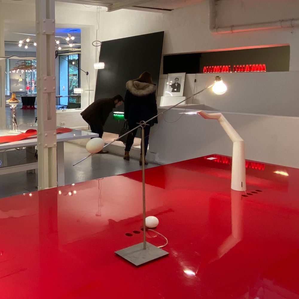 Tischleuchte Ru Ku Ku von Ingo Maurer in einer modernen Ausstellung, mit rotem Tisch und Besuchern.