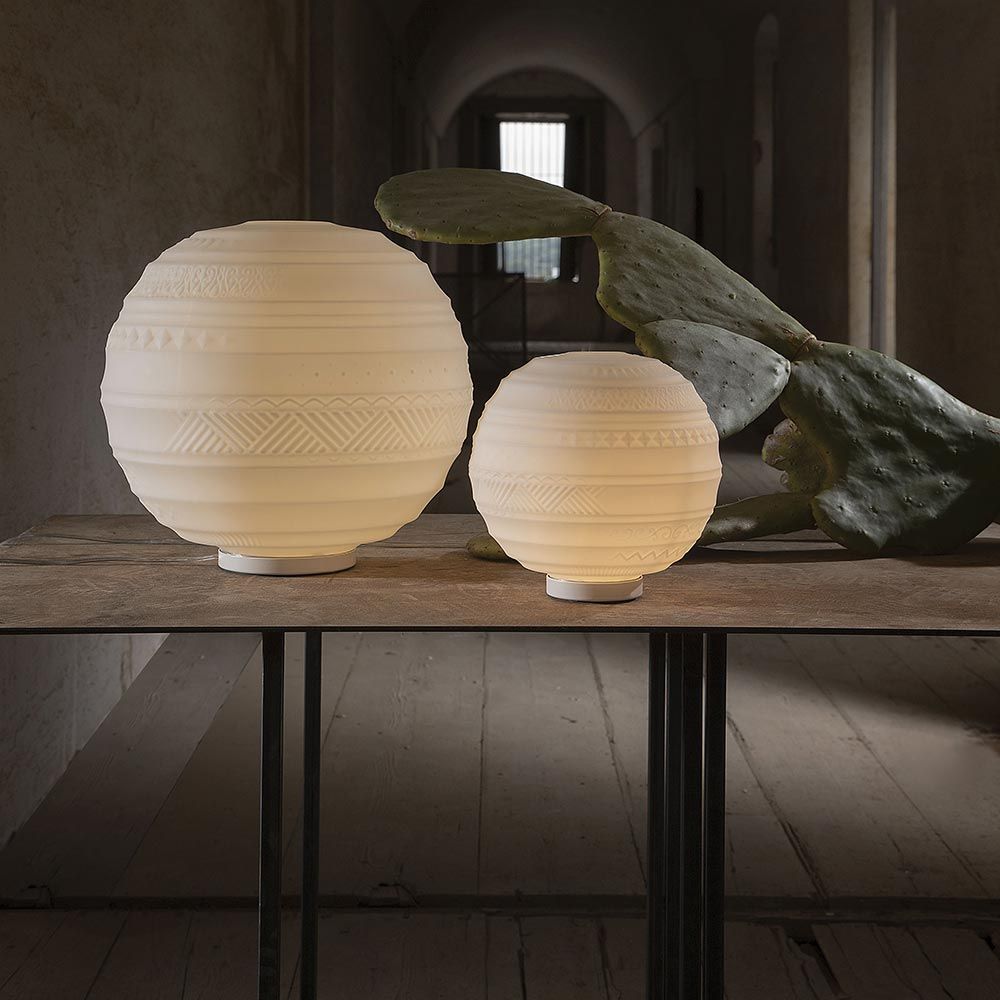 Karman Braille Tischleuchten in Weiß auf Tisch mit Pflanzenblatt, warmes Licht, handgearbeitetes Glasdesign.