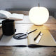 Kundalini LED Akku-Tischlampe Kushi Ø 16cm auf einem Holzschreibtisch mit Büchern und Schreibutensilien.