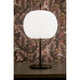 Kundalini Opalglas Tischleuchte Kushi XL 61cm mit Dimmer auf einem Tisch dekoriert vor floralem Hintergrund.