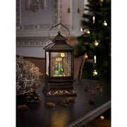 LED Snow lantern with couple small timer water-filled battery-operated on a table with pinecones and Christmas tree in the background.
