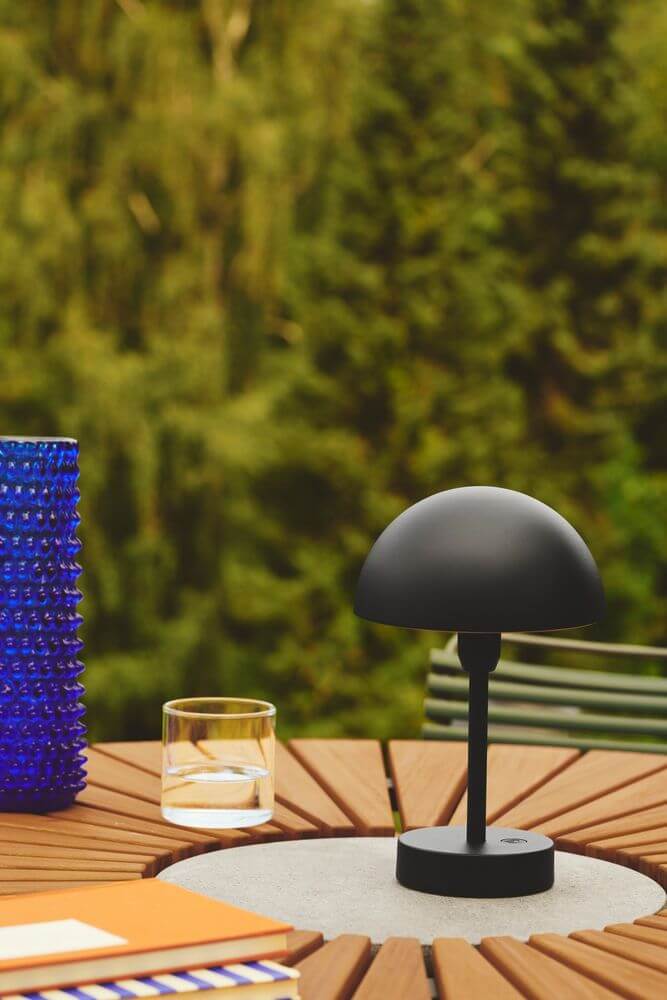 LED Akkuleuchte Ellen To-Go IP44 auf einem Tisch im Freien, umgeben von einem blauen Gefäß und einem Glas.