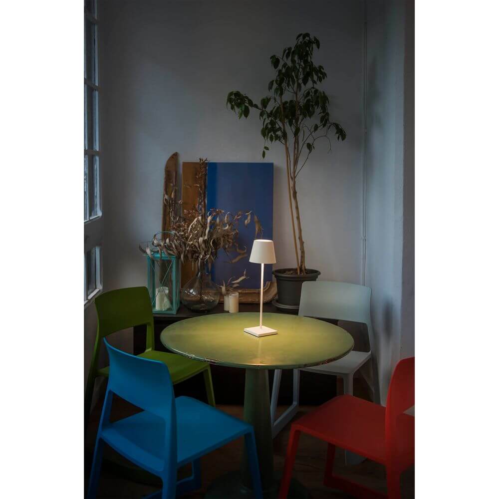 LED Akku-Außentischlampe TOC IP54 Weiß auf einem Tisch in einem modernen Raum mit bunten Stühlen.
