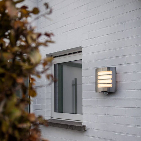 LED Außenwandleuchte Farell mit Sensor IP44 Edelstahl an eine Hauswand montiert, mit warmem Licht und Edelstahl-Rahmen.