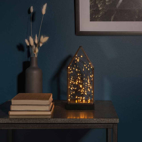 LED Glaslaterne mit schwarzem Holzfundament und 6h Timer Schwarz auf einem Tisch mit Pflanzen und Bücher.