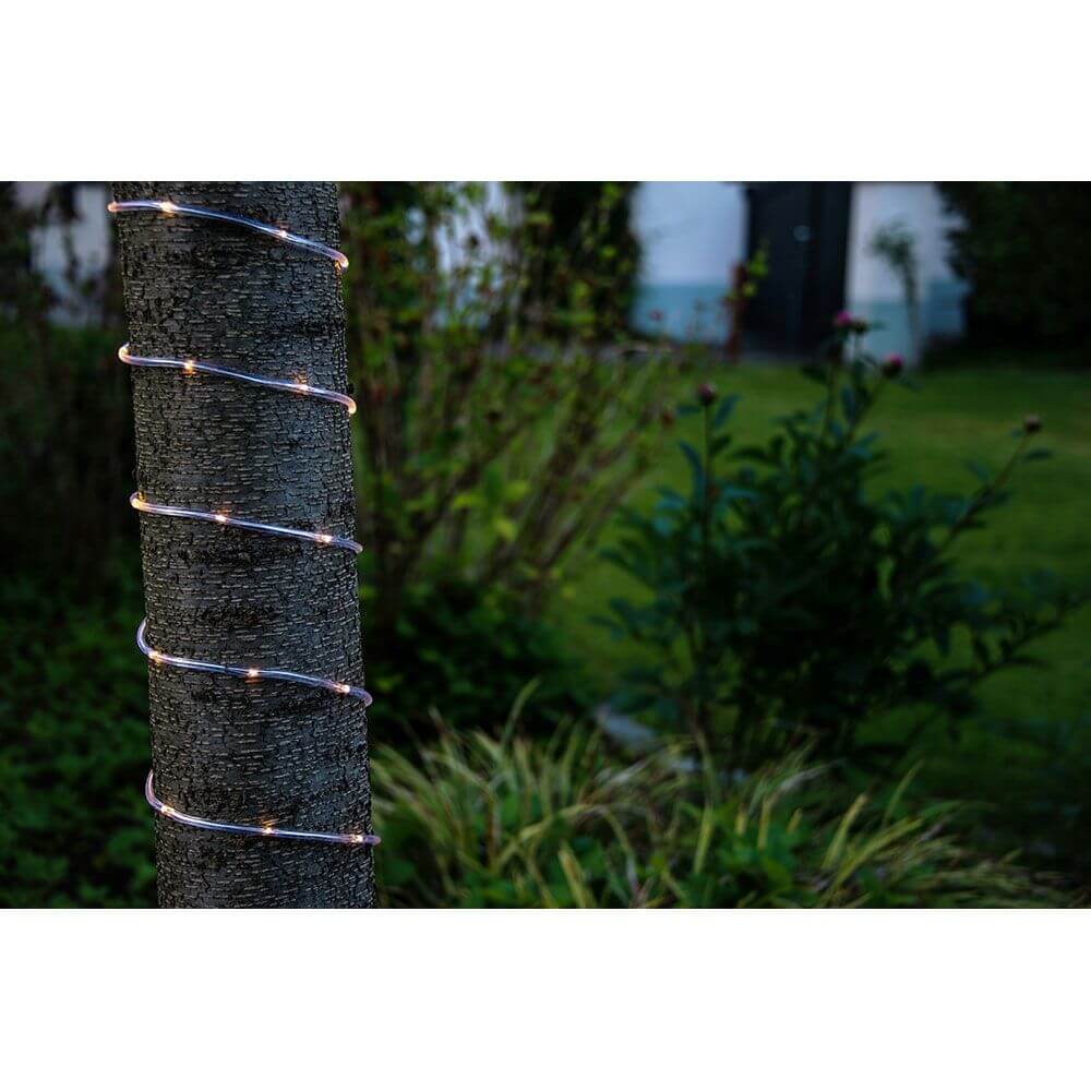 LED Mini Lichterschlauch 10m mit Lichtsensor Timer 130 Warmweiße Dioden batteriebetrieben IP44 um einen Baum gewickelt bei Nacht.