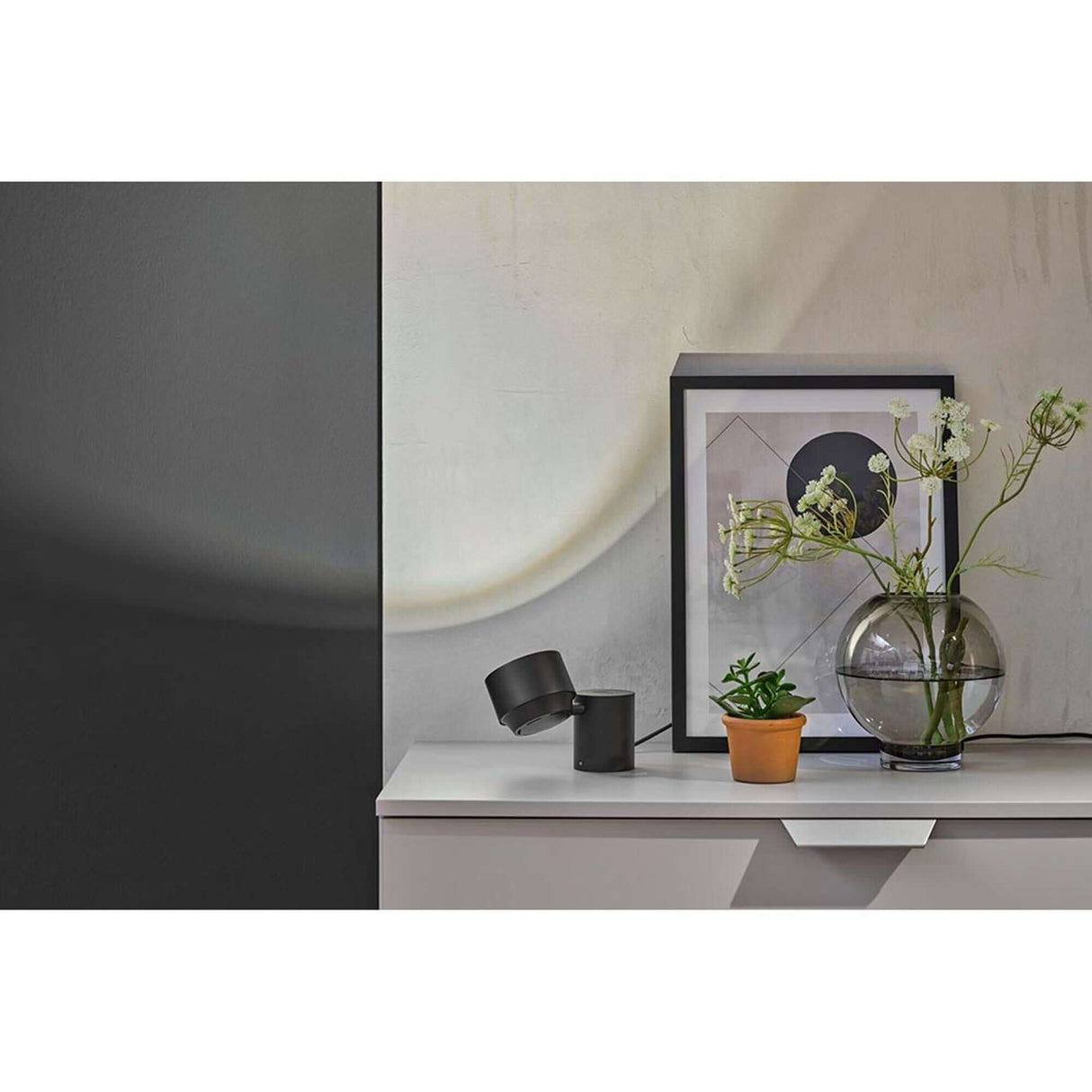 LED Wand- & Tischleuchte Puric Pane mit Touch-Dimmer Schwarz auf einer modernen Kommode mit Pflanzen und Bildern.