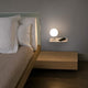 LED Wandleuchte NIKO Links Weiß, Braun neben einem modernen Bett mit Kissen und Mobiltelefon.
