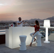 Lámpara de pie de exterior Carmen de 165 cm junto a una zona de bar por la noche con vistas a la costa.