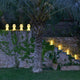 Lola LED-Außen-Tischleuchte Large in einem Garten, umgeben von Pflanzen und stimmungsvollem Licht.