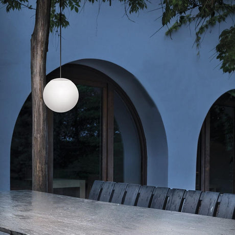 Oh! P65 LED-Pendelleuchte Small über einem Tisch im Freien mit blauem Hintergrund und Bäumen.