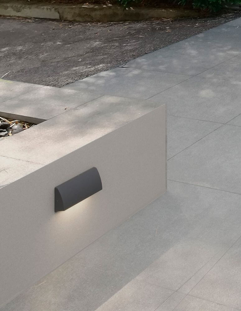 Liv LED Außenwandleuchte 3000K IP54 – dunkle moderne Außenwandleuchte an hellgrauer Mauer, warmes Licht nach unten strahlend
