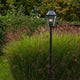 Lutec London Außen-Standleuchte IP44 300lm in einem Garten mit üppigem Grün und sanften Licht.