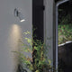 Modena Energiespar Wandspot schwenkbar Grau, klares Glas an einer Wand mit Pflanze davor.