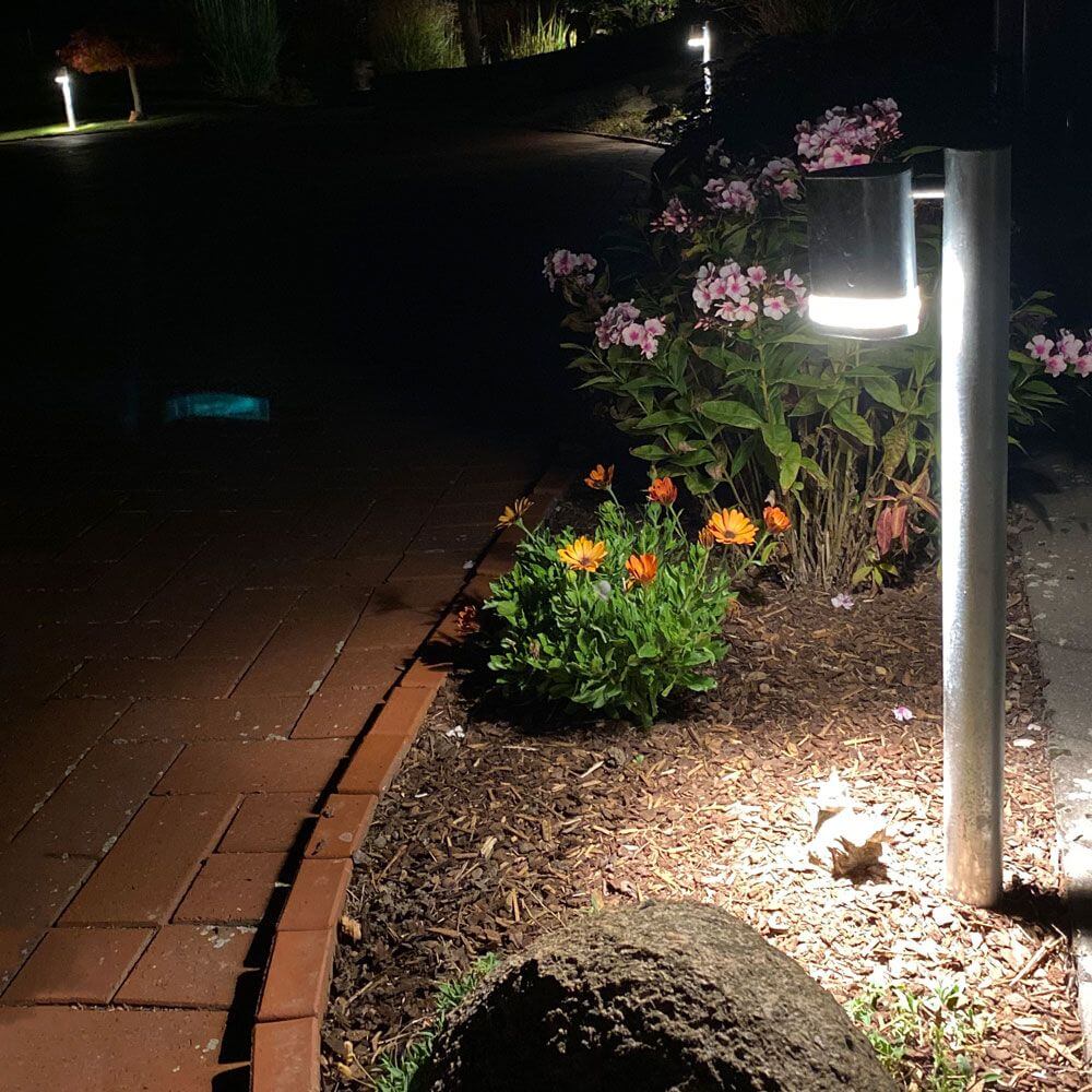 Modena Wegeleuchte mit Pfahl klares Acrylglas, Reflektor neben blühenden Pflanzen in der Dunkelheit.
