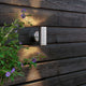 Monza LED lampa ścienna zewnętrzna z podwójnym stożkiem światła na ścianie drewnianej z roślinami na pierwszym planie.