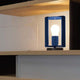 Nemo Pivotante À Poser Tischlampe drehbar auf einem Regal mit blauer Struktur und leuchtender Glühbirne.