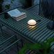 Bring LED Akkuleuchte Ø 12cm IP54 auf einem Tisch im Freien, umgeben von Pflanzen und einem Buch.
