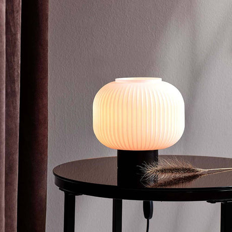 Tischleuchte Milford Schwarz, Opalweiß auf einem Tisch mit warmem Licht und eleganter Form.