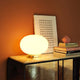 Oluce Glas Tischlampe Alba Alu, Weiß auf einem Tisch mit Bücher und Kunstwerk im Hintergrund.
