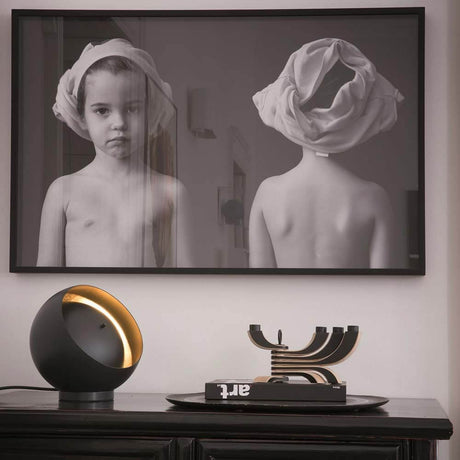 Oluce LED Tischleuchte Eva Schwarz, Goldfarben auf einem Tisch mit einem Kunstwerk im Hintergrund.