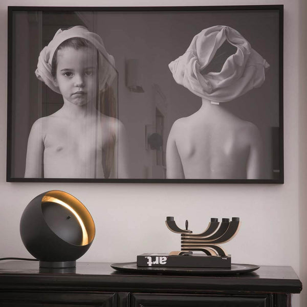 Oluce LED Tischleuchte Eva Schwarz, Goldfarben auf einem Tisch mit einem Kunstwerk im Hintergrund.