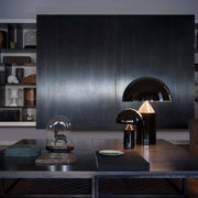 Oluce bordlampe Atollo 70 svart på et elegant bord med moderne møbler i bakgrunnen.
