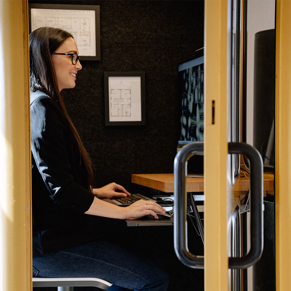 Frau arbeitet an einem Computer in einem modernen Arbeitsraum, umgeben von Plänen und strukturierten Wänden.