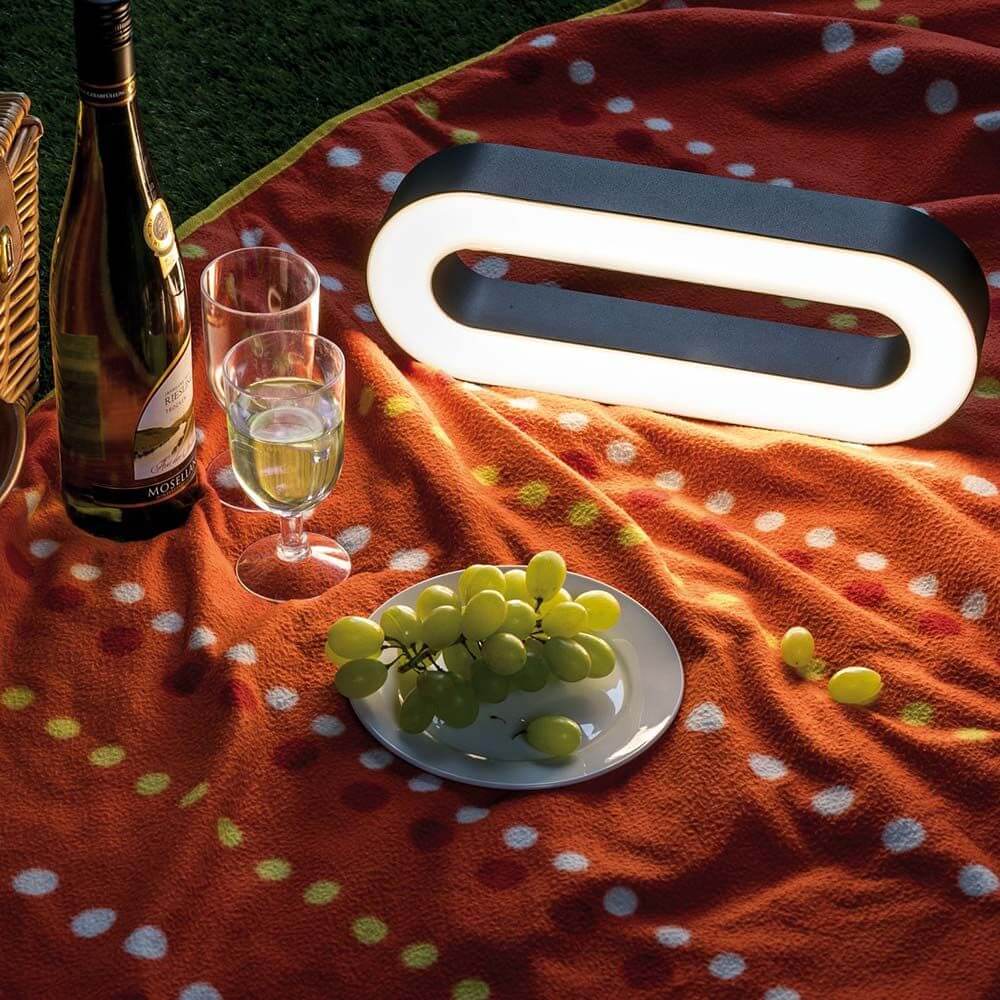 LED Akkuleuchte Moval IP44 370lm 3000K USB auf Picknickdecke mit Wein und Trauben.