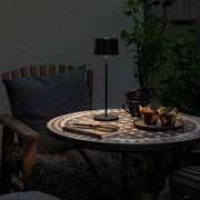 Stylische Positano Outdoor LED Akku Tischleuchte auf einem Tisch im Freien, umgeben von Snacks und einem gemütlichen Ambiente.