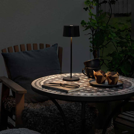 Stylische Positano Outdoor LED Akku Tischleuchte auf einem Tisch im Freien, umgeben von Snacks und einem gemütlichen Ambiente.